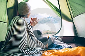 Young woman covered with warm plaid meets cold morning in sitting in a touristic camping tent with a cup of hot tea. Romantic cam