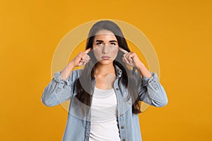 Young woman covering ears with fingers on yellow background