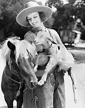 Young woman in a cowboy hat holding a goat while leaning against her pony