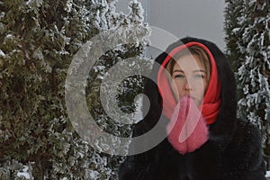 Young woman in a cozy fur coat with a hood and pink mittens on the street in winter. Portrait of a girl dressed in winter clothes