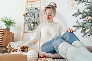 Young woman in cozy sweater petting her cat while decorating potted Christmas tree in light modern Scandinavian interior home. Eco