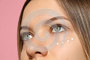 Young woman with cream around eye on pink background, closeup