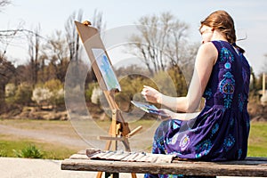 Young woman creative painter draws brush watercolor paints on canvas outdoors. Spring inspiration.