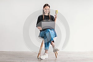 Young woman with credit card and laptop sitting on chair, buying goods on internet. Online shopping, remote banking