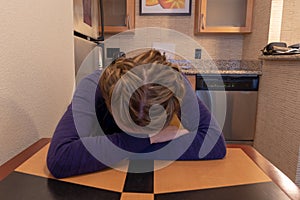 Young woman cries and buries her head and face in her arms while sitting at a kitchen table alone