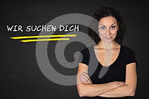 Young woman with crossed arms and `Wir suchen dich` text on a blackboard background. Translation: `We are looking for you` photo