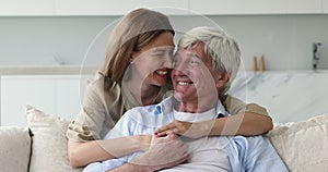 Young woman cuddling her aged daddy sits on sofa