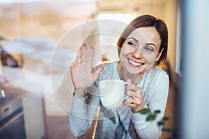 A young woman with cup of coffee looking out of a window, waving goodbye.