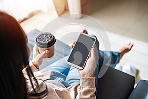 Young woman with cup of coffee using mobile phone and relaxing on sofa in living room at cozy home