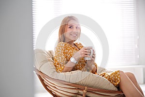 Young woman with cup of drink relaxing in papasan chair near window