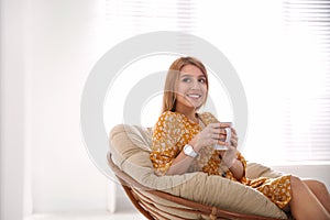 Young woman with cup of drink relaxing in papasan chair near window. Space for text