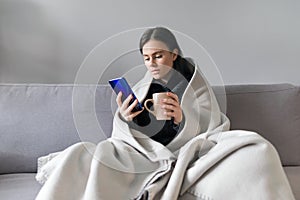 Young woman with cup, smartphone, sitting at home on sofa under warm fleece blanket