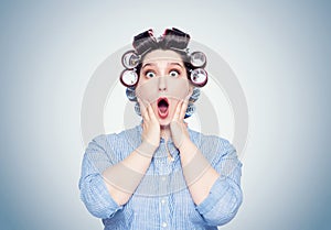 Young woman with curlers opening her mouth holds her hands behind her head, very surprised, scared