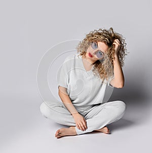 Young woman with curly blonde hair in white pants and blouse sits on the floor holding hand at her tilted head