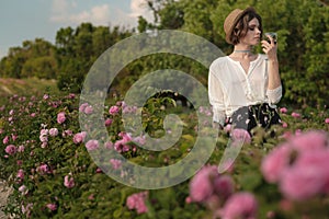 Young woman with curly hair posing near roses in a garden. The concept of perfume advertising