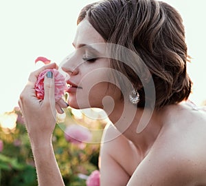Young woman with curly hair posing near roses in a garden. The concept of perfume advertising