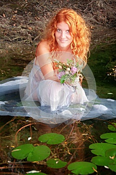 Young woman with curly red hair is sitting joyfully, with white dress happy in the water in the lake