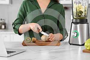 Young woman cutting kiwi for smoothie at white table in kitchen, closeup