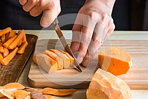 Young woman cutting with knife sweet potato into wedges, peels on wood table, sliced carrots