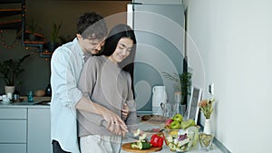 Young woman cutting vegetables cooking salad then kissing and hugging husband in kitchen in apartment