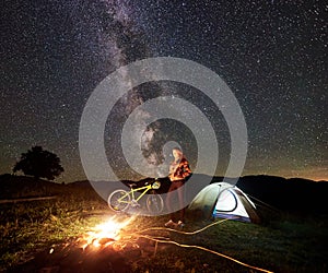 Woman resting at night camping near campfire, tourist tent, bicycle under evening sky full of stars