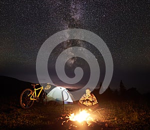 Woman resting at night camping near campfire, tourist tent, bicycle under evening sky full of stars