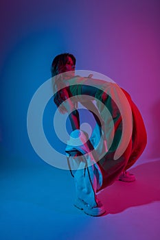 Young woman dancer in stylish youth clothes in gym shoes dancing and posing in a room with bright neon blue-pink color.Attractive