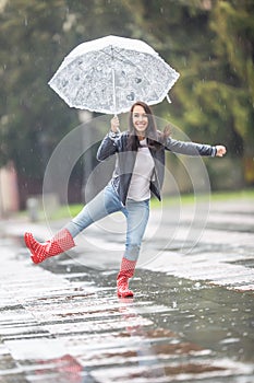 Young woman dances in the rain in the park, holding an umbrella, wearing rainboots