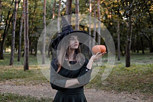 Young woman in dark dress and witch hat holds a pumpkin in her hands. Halloween party costume. Forest, park with autumn