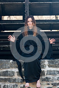 Young Woman with dark long hair in black robes in front of an Old Wooden Water Mill. Witches. Halloween concept. Witchcraft