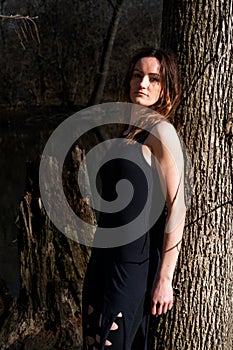 Young woman with dark long hair in black robes is leaning against the tree in the middle of the forest. Back to Nature concept