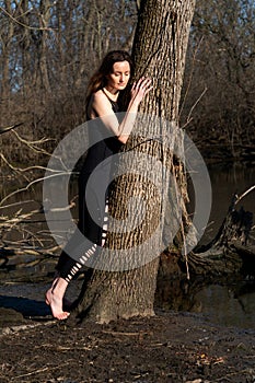 Young woman with dark long hair in black robes is leaning against the tree in the middle of the forest. Back to Nature concept