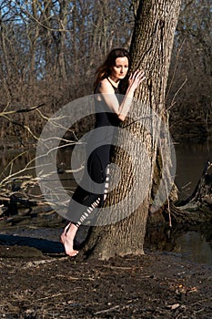 Young woman with dark long hair in black robes is leaning against the tree in the middle of the forest. Back to Nature concept