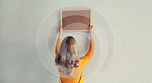 Young woman decorating interior, hanging blank a photo frame mockup on white wall in a new house