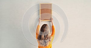 Young woman decorating interior, hanging blank a photo frame mockup on white wall in a new house