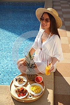 Young woman with delicious breakfast on tray near swimming pool