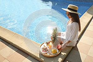 Young woman with delicious breakfast on tray near swimming pool. Space for text