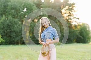 Young woman in a denim shirt and pink skirt holds a hat in her hands, she smiles and walks in nature in the summer