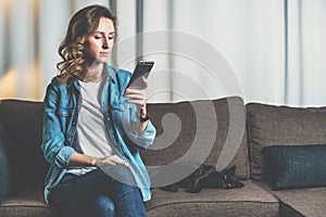 Young woman in denim shirt sitting at home on couch and using smartphone,nearby lies cat. Girl uses digital gadget.