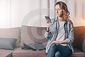 Young woman in denim shirt sitting at home on couch and using smartphone,nearby lies cat. Girl uses digital gadget.