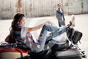 Young woman in denim vest sitting on motorcycle and stylish man in sunglasses standing behind