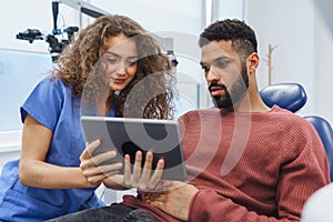 Young woman dentist doing preventive examination to multiracial man, showing him x-ray image in digital tablet.