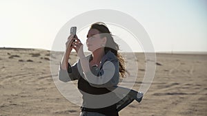 Young woman in desert taking photos on mobile phone. Sand storm, strong wind.