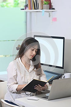 Young woman designer working with laptop computer and reading information on notebook at creative home office.