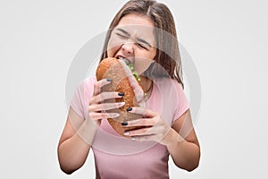 Young woman devour sandwich. She hold it in hands and keep eyes closed. Isolated on grey background.