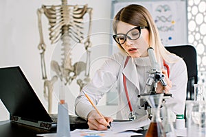 Young woman doctor, biochemist, scientist looking through a microscope in a laboratory, making notes on the papers and