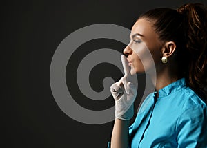 Young woman doctor in blue medical gown and latex gloves holds finger at her lips gesturing shh sign calling for silence