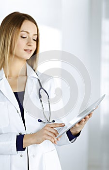 Young woman-doctor is holding a tablet computer in her hands, while standing in a clinic. Portrait of friendly female