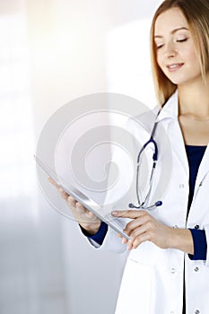 Young woman-doctor is holding a tablet computer in her hands, while standing in a sunny clinic. Portrait of friendly