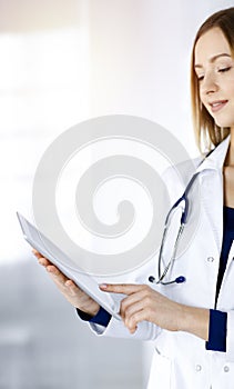 Young woman-doctor is holding a tablet computer in her hands, while standing in a sunny clinic. Portrait of friendly